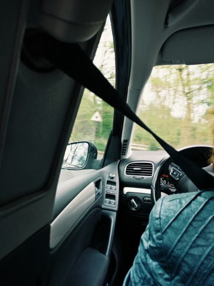 View across a vehicle cabin from the passenger side, with the seatbelt running diagonally past the door pillar towards the dashboard and open road ahead.