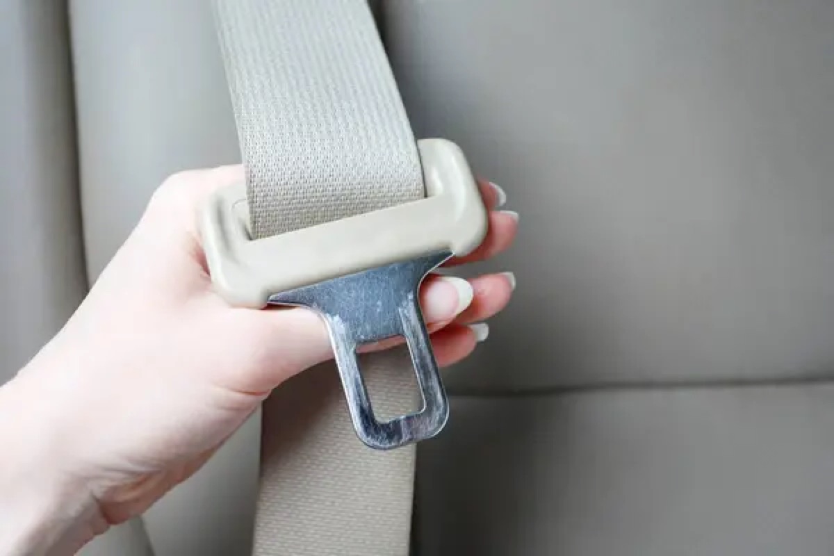 A hand holding a beige seatbelt latch plate and matching webbing in front of a beige vehicle seat.