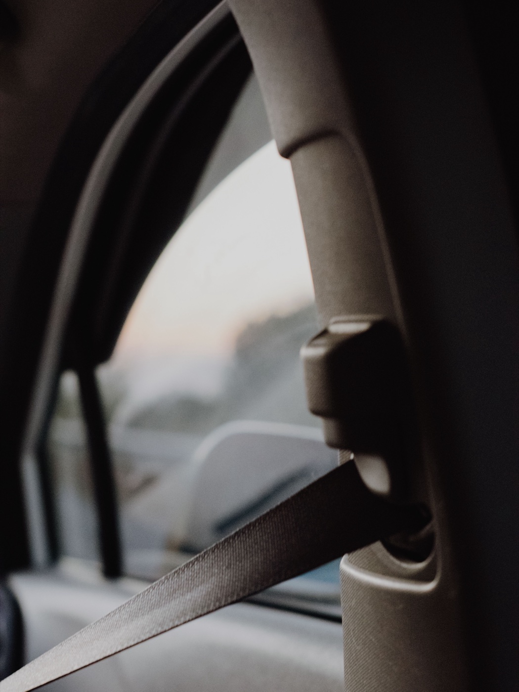 Seatbelt webbing running down a vehicle door pillar past a height adjustment fitting, seen against evening light through the window.