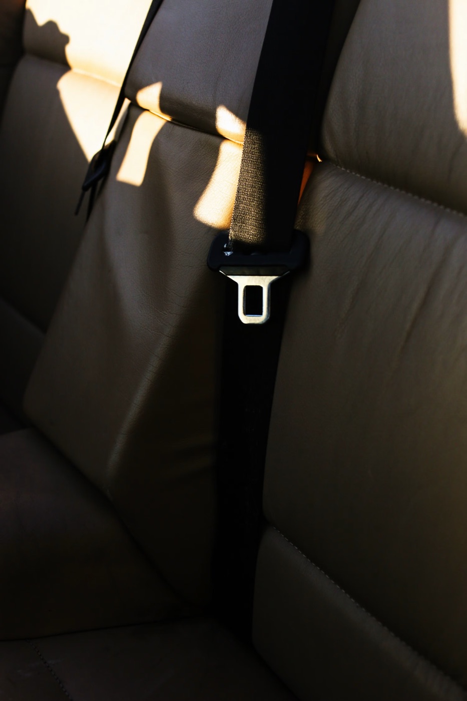 A black seatbelt buckled across a pale rear bench seat, lit by low sunlight through the rear window.